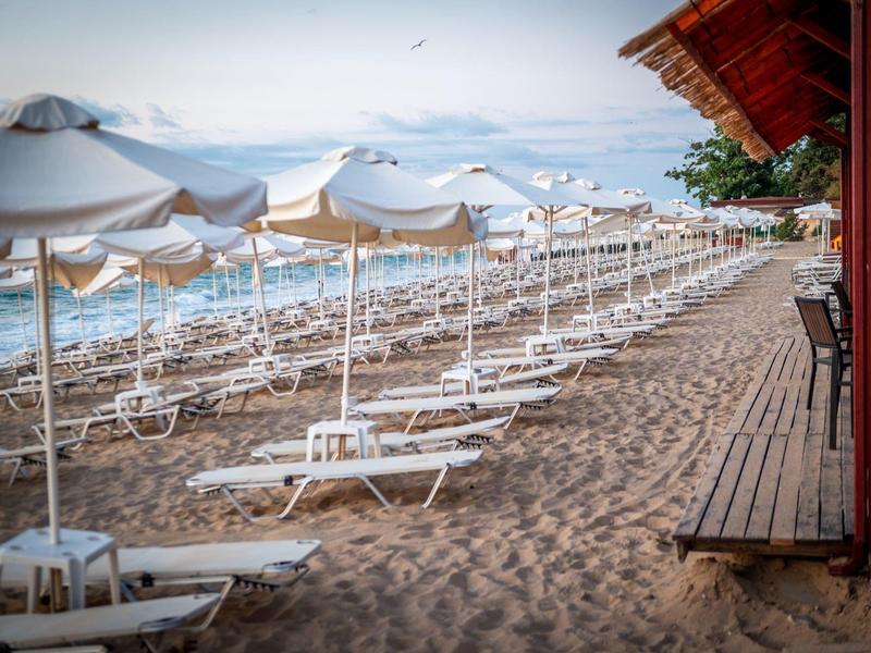 Prázdná plážová lehátka a slunečníky na klidné písečné pláži vedle dřevěné promenády a červené budovy.