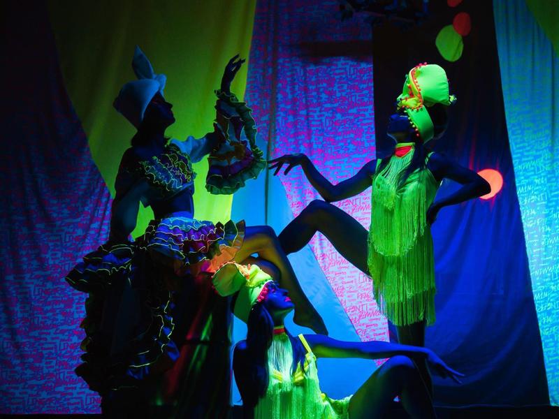 Dva tanečníci v barevných kostýmech na živém pozadí s dramatickým osvětlením.