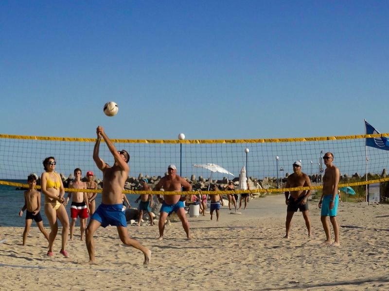 Persone che giocano a beach volley al sole su un campo di sabbia con cielo azzurro.