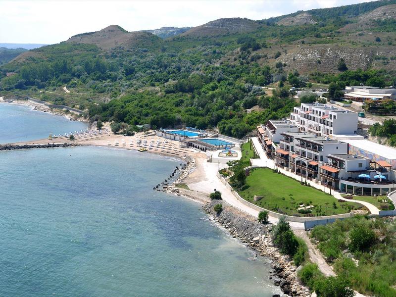 Complesso alberghiero sulla riva con piscina, ombrelloni e colline boschive sullo sfondo.