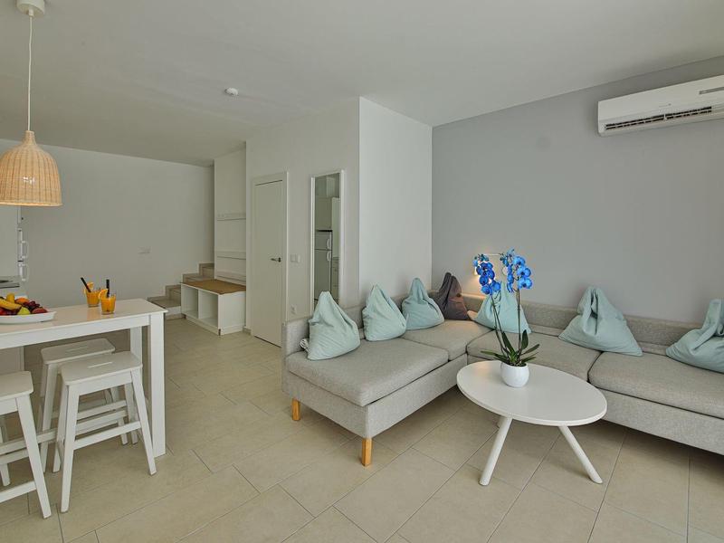 Zona giorno moderna con divano ad angolo grigio, tavoli bianchi e cucina aperta con zona pranzo.