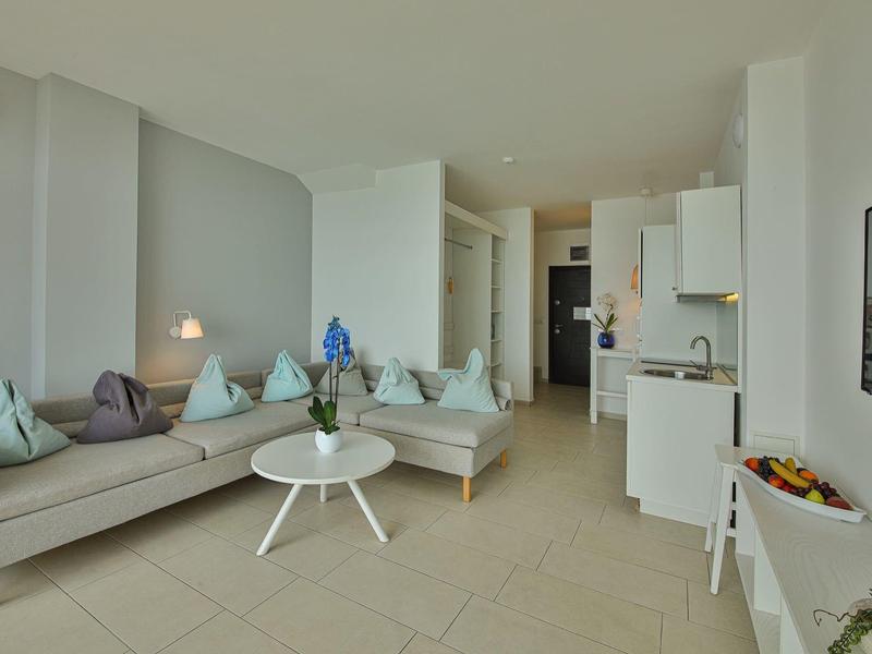 Soggiorno luminoso con divano, cuscini, tavolino rotondo e cucina aperta in un appartamento per vacanze.