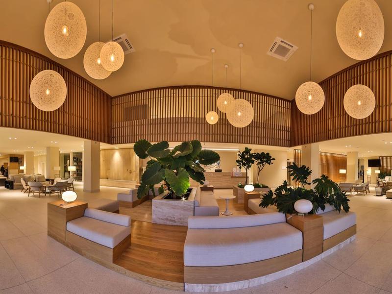 Luminosa hall d'hotel con aree salotto moderne, piante e luci rotonde a soffitto.