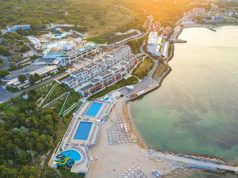 Vista aerea di un resort sulla spiaggia con piscine, lettini e costa limpida.