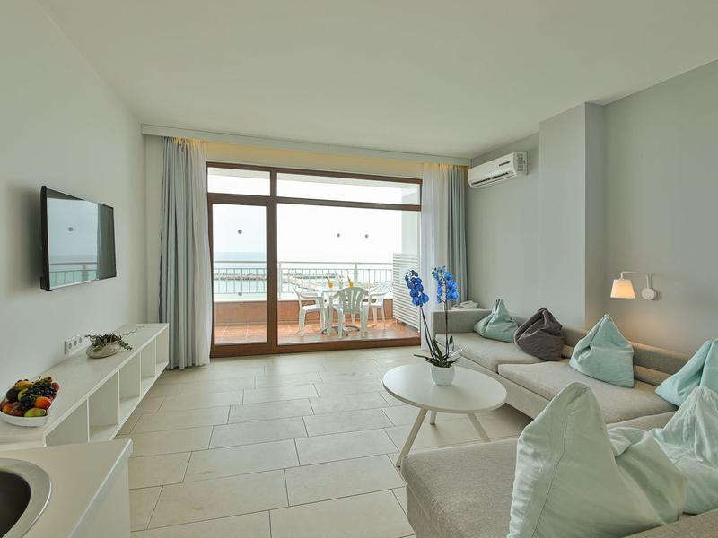 Soggiorno luminoso con divano, tavolino, TV e accesso al balcone con vista mare.