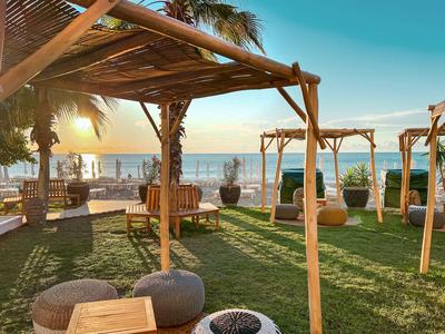 Bar de playa soleado con muebles de madera, toldos de paja y zonas de descanso junto al mar.