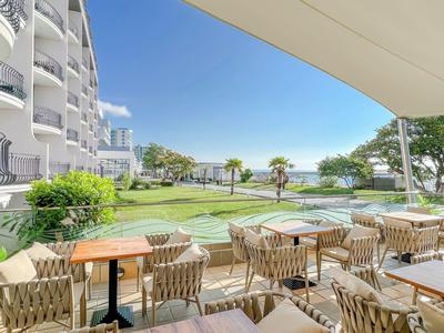 Terrasse avec chaises et tables donnant sur une pelouse, des palmiers et la mer par temps ensoleillé