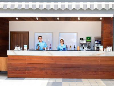 Bar d'hôtel avec deux employés derrière un grand comptoir en bois avec machines à café et boissons.