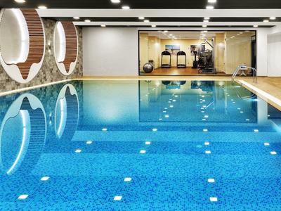 Piscine intérieure moderne avec sol en bois, tabourets et salle de sport en arrière-plan.