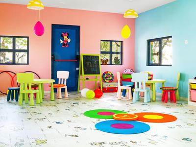 Salle de jeux colorée avec petites tables, chaises, jouets et fenêtres laissant entrer la lumière du jour.