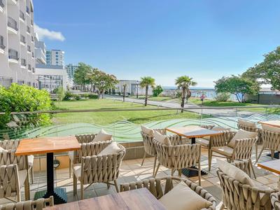 Terraza con mesas y sillas frente a un hotel con vista a un jardín y al mar.