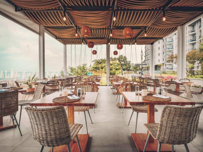 Terrasse extérieure couverte avec toit en bois et chaises en osier offrant une vue sur un parc et des bâtiments.