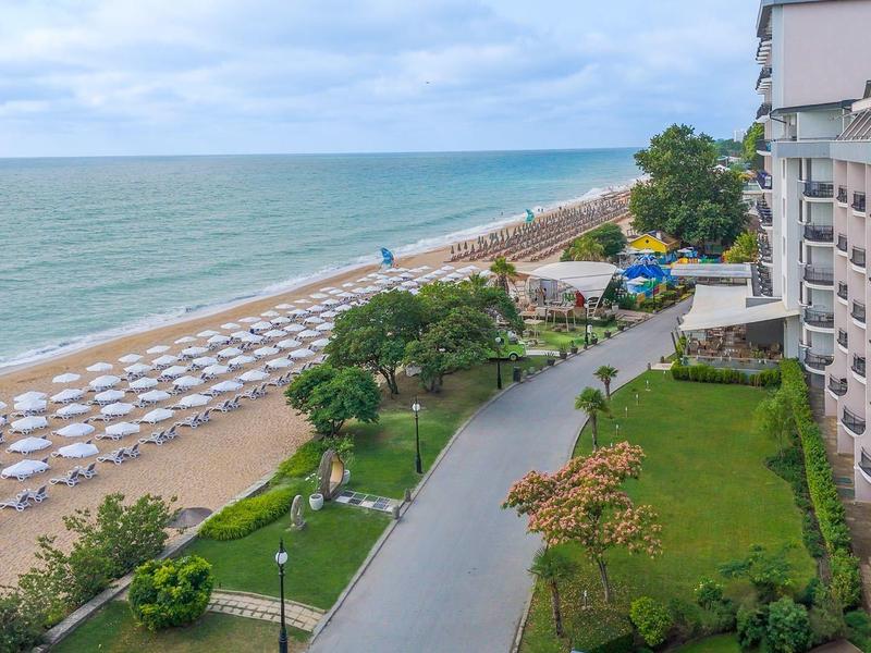 Vue sur une plage avec des chaises longues, un jardin adjacent et un bâtiment d'hôtel à plusieurs étages.