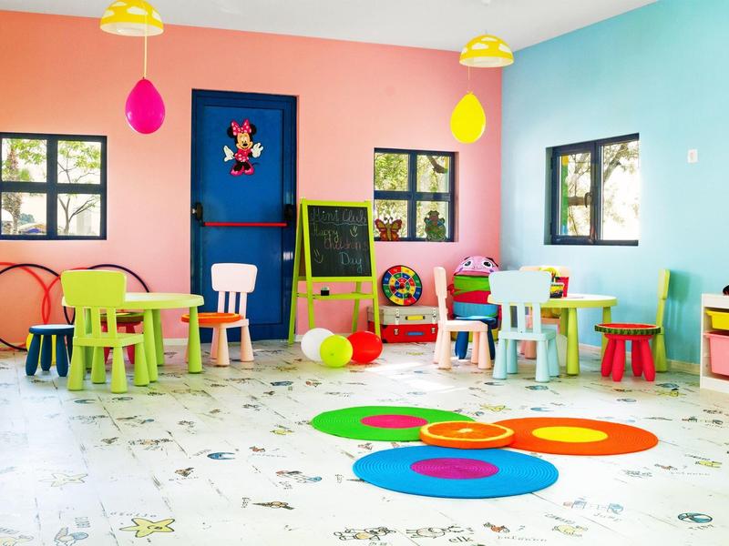Salle de jeux colorée avec petites tables, chaises, jouets et fenêtres laissant entrer la lumière du jour.