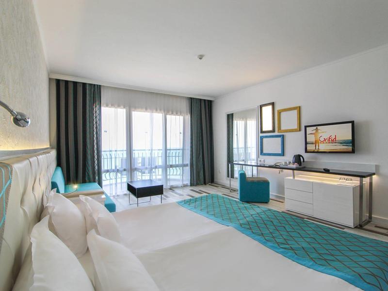 Chambre d'hôtel moderne avec grand lit, balcon et vue sur la mer, décor lumineux.