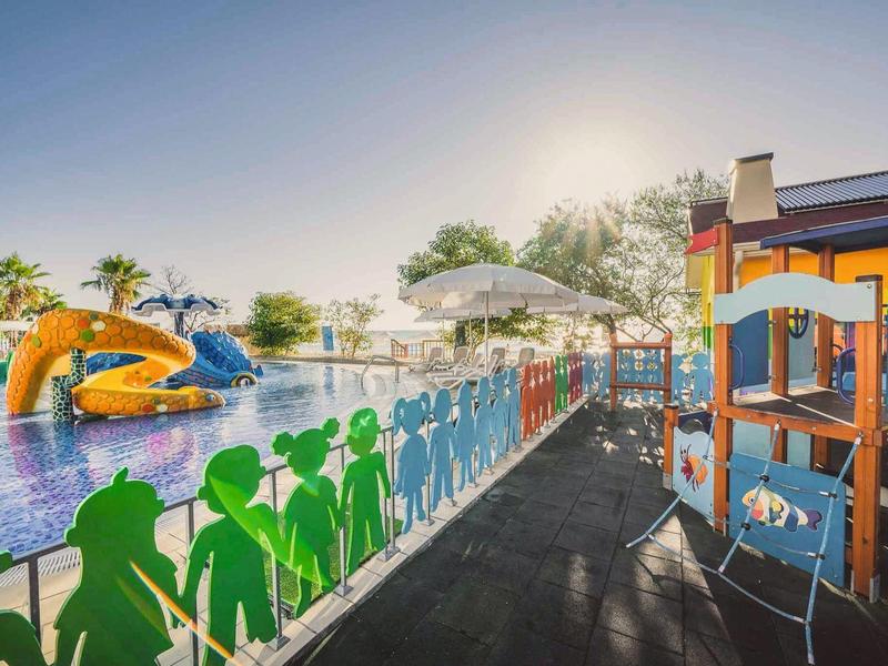 Aire de jeux pour enfants à côté d'une piscine avec toboggan aquatique et parasols par temps ensoleillé.