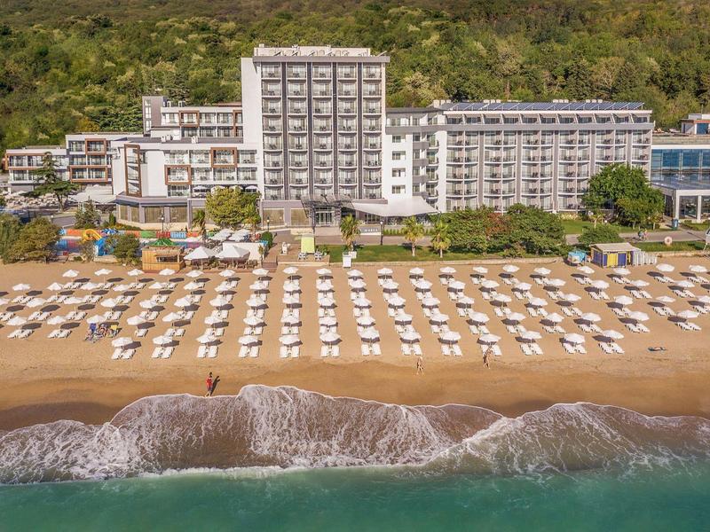 Grand hôtel à plusieurs étages sur la plage avec de nombreux parasols blancs et chaises longues devant.