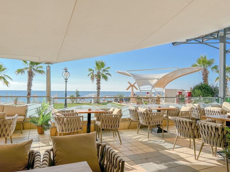 Terrasse ouverte avec tables et chaises, vue sur la mer et palmiers par temps ensoleillé.
