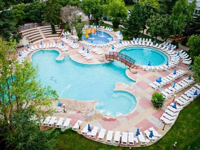 Großer Hotelpool mit Liegestühlen, Sonnenschirmen und umliegendem Grünbereich.