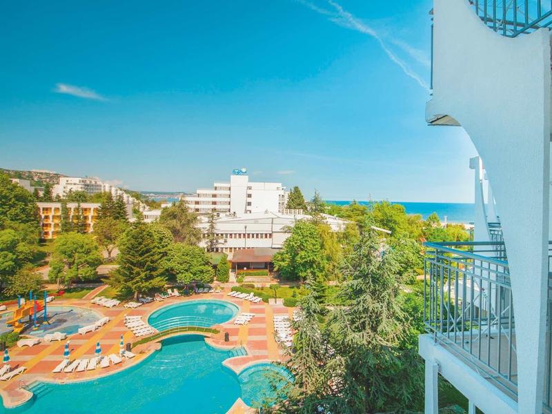 Uitzicht vanaf balkon op hotelzwembad, tuin en zee onder heldere blauwe lucht.