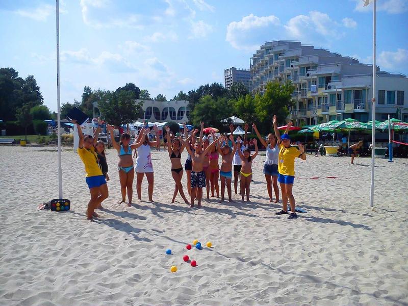 Groep mensen en entertainers op het strand voor een hotel op een zonnige dag.