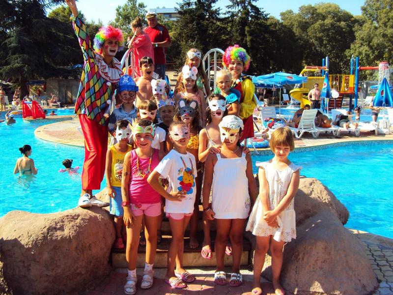 Kinderen poseren met twee clowns naast een buitenzwembad op een zonnige dag.