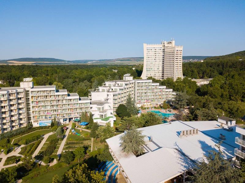 Groot hotelcomplex met meerdere gebouwen omringd door bomen en beboste heuvels onder een blauwe lucht.