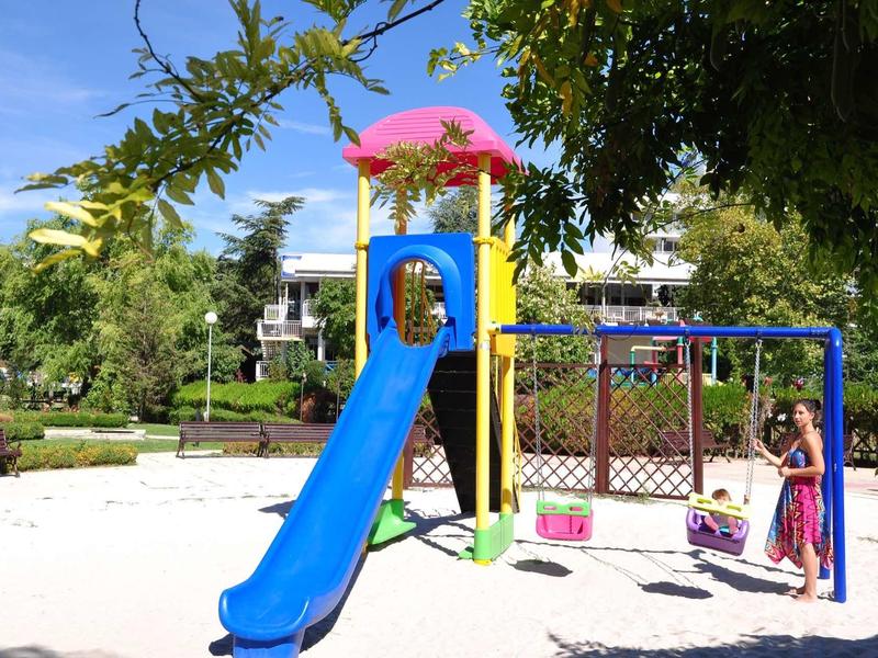 Kinderspeeltuin met blauwe glijbaan en schommel in een zonnige tuin voor een hotelgebouw.