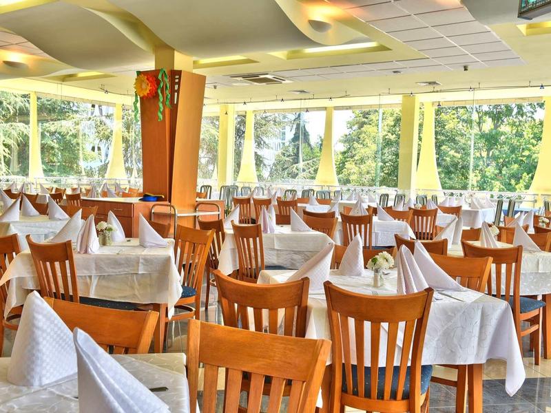 Lichte eetzaal met meerdere houten tafels en stoelen, witte tafelkleden, grote ramen met uitzicht op groene bomen.