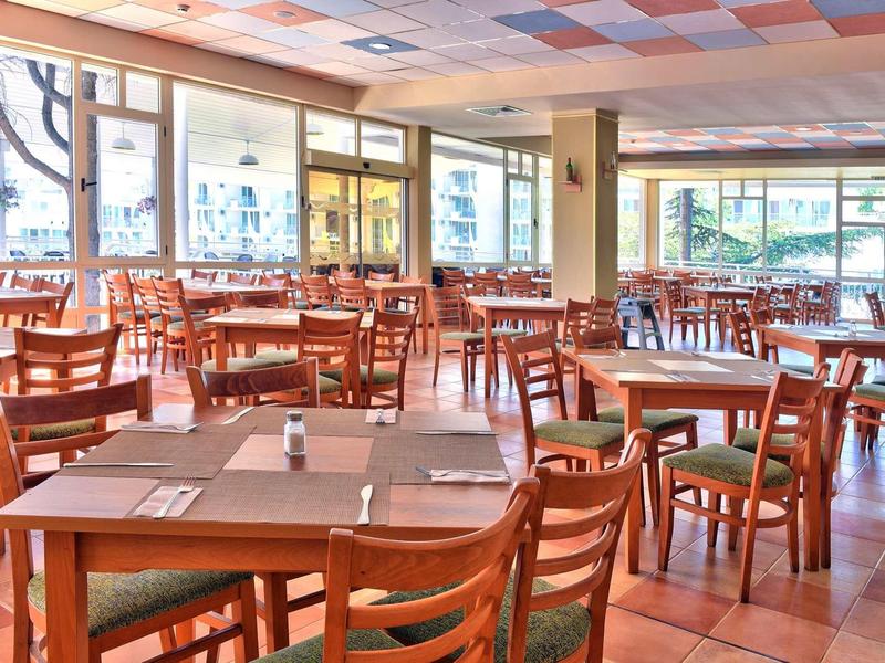 Licht, ruim restaurant met houten tafels en stoelen, grote ramen en betegelde vloer.