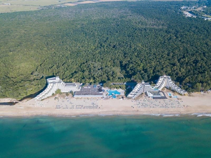 Luchtfoto van een hotel aan het strand met zwembad, omgeven door bos en helder water.