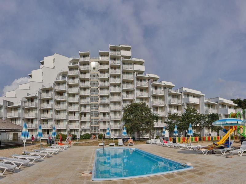 Meerlagig hotel met balkonkamers naast een zwembad en ligstoelen onder een blauwe hemel.