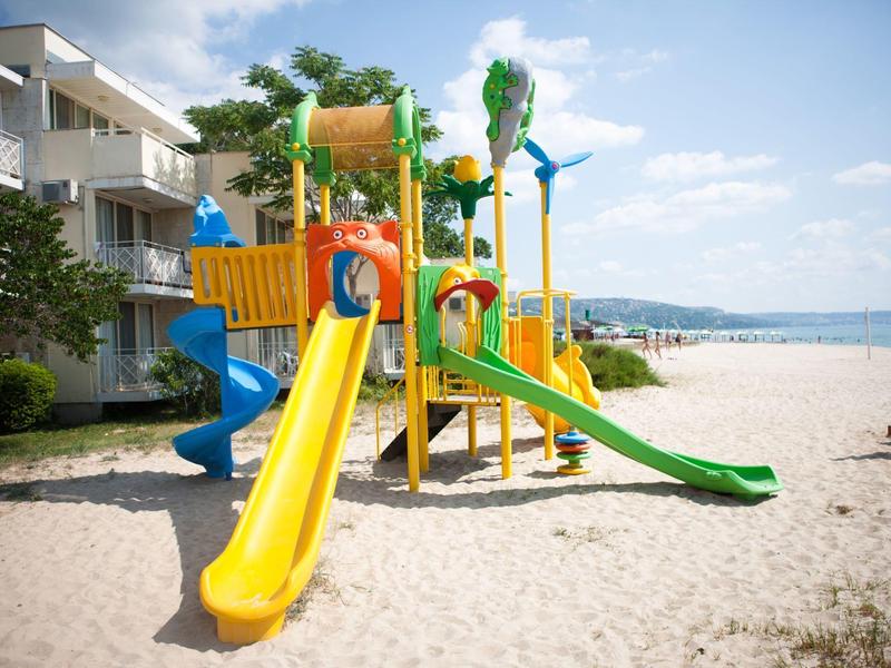 Kleurrijke kinderspeelplaats met glijbanen op zandstrand naast hotelgebouw.