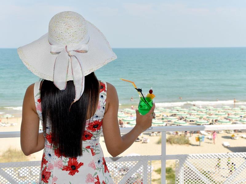 Vrouw met hoed en cocktail kijkt uit op het strand en de zee.