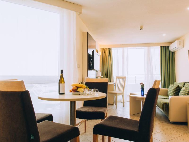 Lichte hotelkamer met eetgedeelte, bank, tv en balkonzicht.