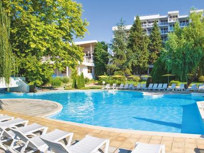 Una piscina redonda de hotel con tumbonas, rodeada de árboles y un edificio de varios pisos al fondo.