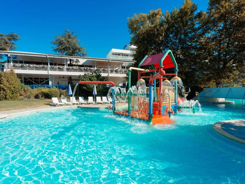 Piscina para niños con estructura de juego colorida frente a un edificio de hotel bajo cielo azul.