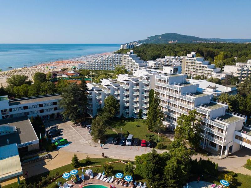 Complejo hotelero con varios edificios blancos junto a la playa y el mar, rodeado de árboles y montañas al fondo.