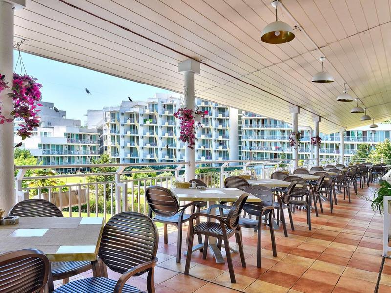 Terraza cubierta con mesas y sillas, al fondo apartamentos modernos y área de jardín.
