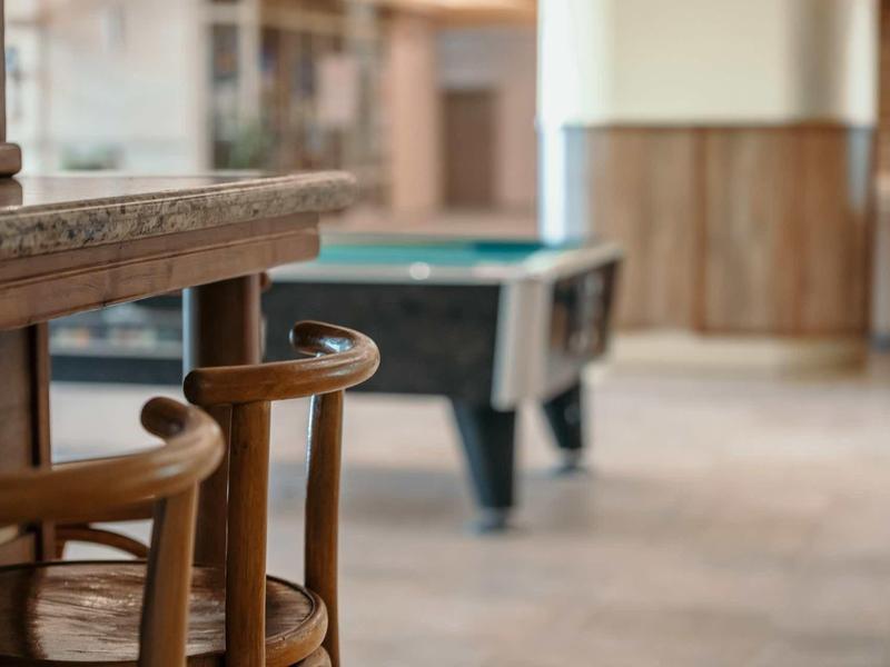 Sillas de madera frente a una mesa con una mesa de juego desenfocada al fondo en una sala común.