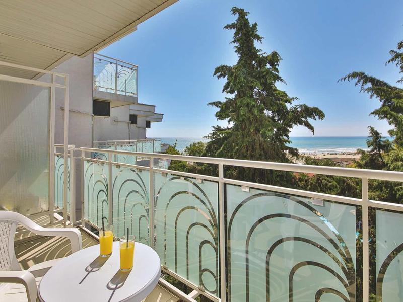 Balcón con mesa y sillas, vista al mar y árboles bajo un cielo azul despejado.