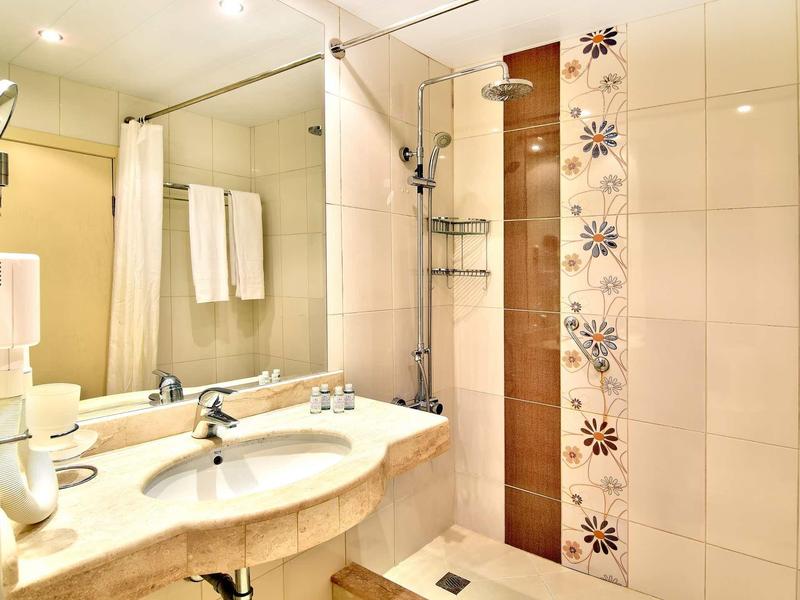 Baño de hotel luminoso con doble lavabo, espejo de pared y ducha tipo walk-in con cortina.