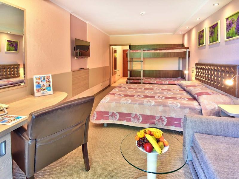 Habitación de hotel con dos camas, literas, escritorio, sillón y mesa con frutas.