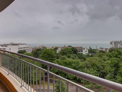 Balcony with gray railing overlooking green trees and buildings by the sea under cloudy sky.