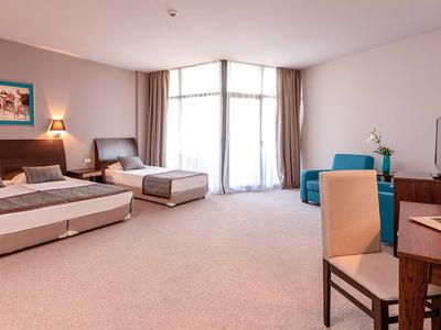 Prostorný hotelový pokoj se dvěma postelemi, stolem a velkými okny s záclonami.