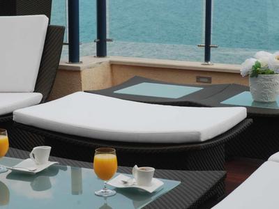White lounge chairs and glass tables with drinks next to a water view on a terrace.