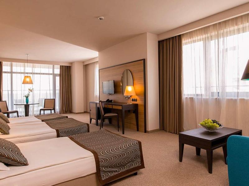 Světý hotelový pokoj se dvěma jednolůžky, psacím stolem, křeslem a velkým oknem s závěsy.