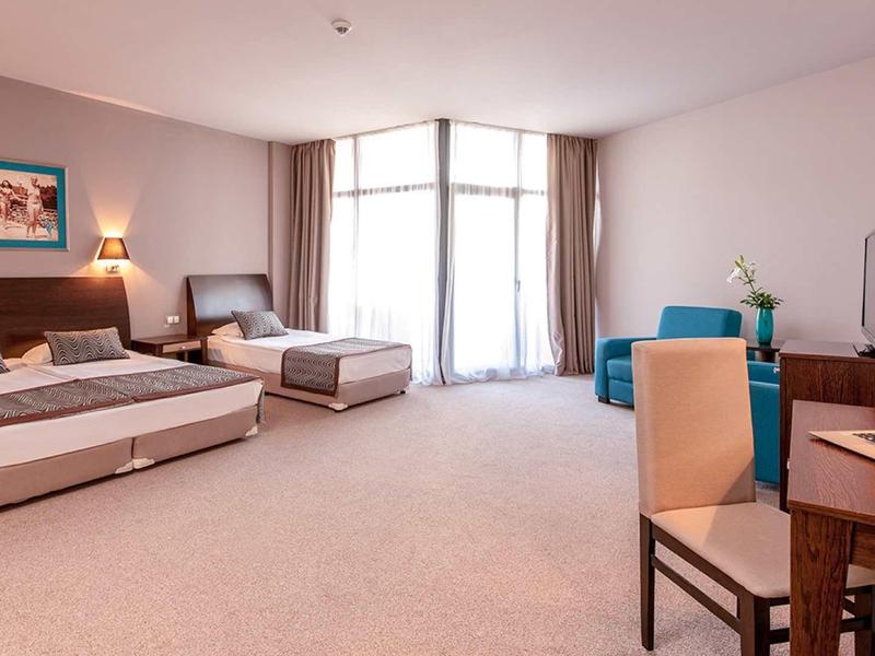 Prostorný hotelový pokoj se dvěma postelemi, stolem a velkými okny s záclonami.