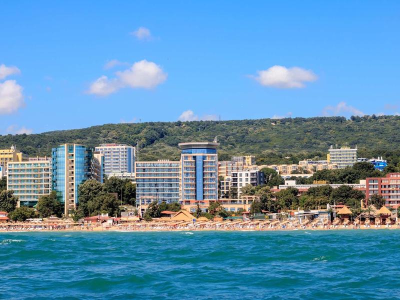 Pobřeží s hotely a zelenými kopci pod jasnou modrou oblohou a klidným mořem.