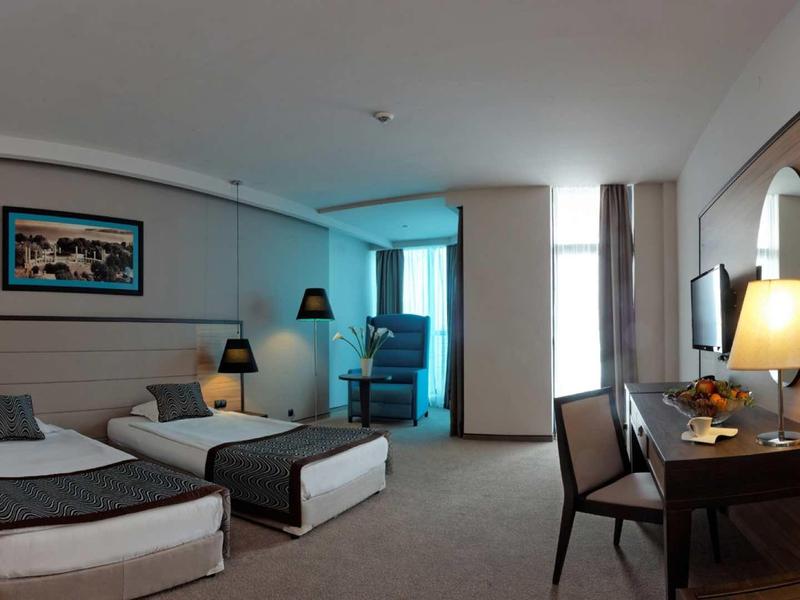 Moderní hotelový pokoj se dvěma jednolůžky, psacím stolem a posezením u okna.